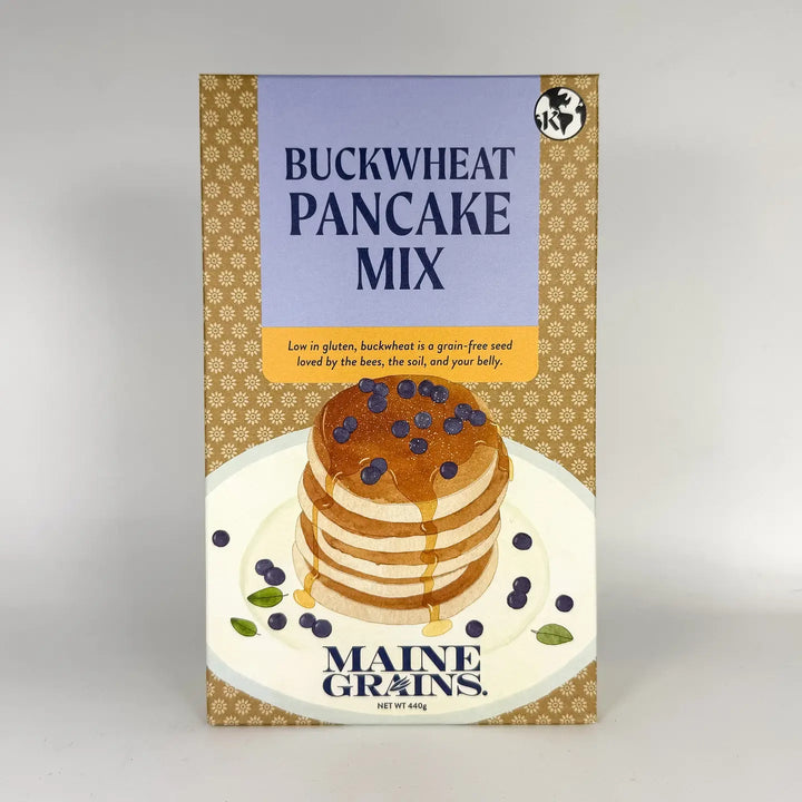 Box of Maine Grains Buckwheat Pancake Mix with a stack of pancakes and blueberries on a white plate.